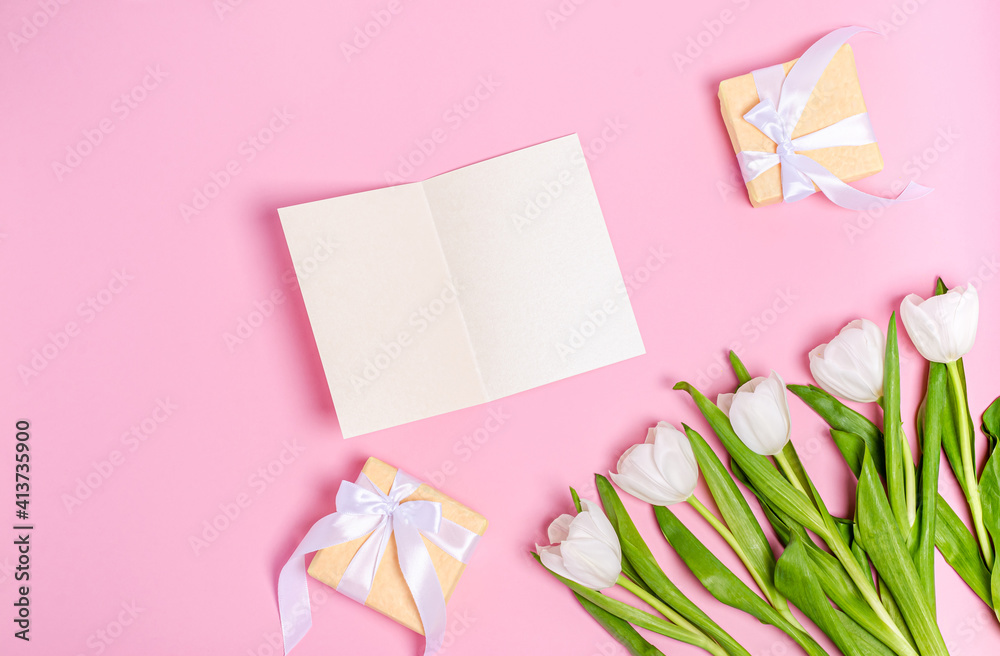 Bouquet of white tulips with a greeting card and boxes for gifts on a pink background.