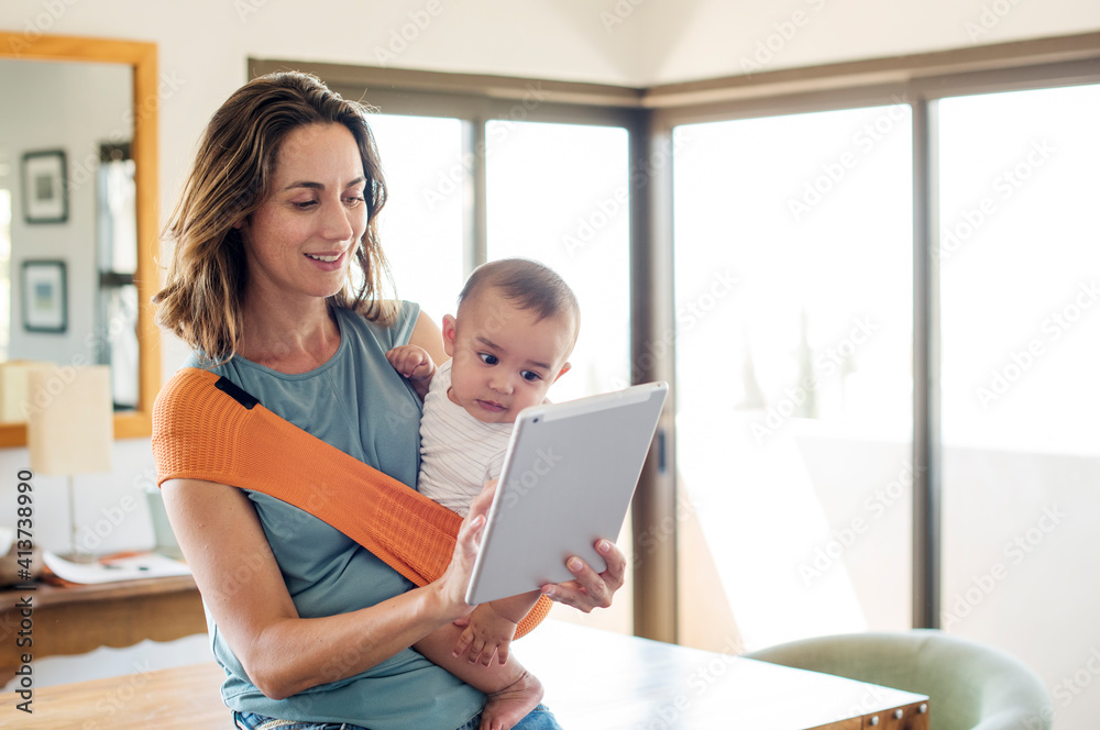 Mother carrying baby boy in carrier while using tablet computer at home Mother carrying baby boy in carrier while using tablet computer at home