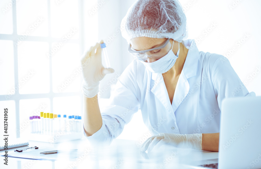 Professional female scientist in protective eyeglasses researching tube with reagents in sunny laboratory toned in blue. Medicine and science researching Professional female scientist in protective eyeglasses researching tube with reagents in sunny laboratory toned in blue. Medicine and science researching