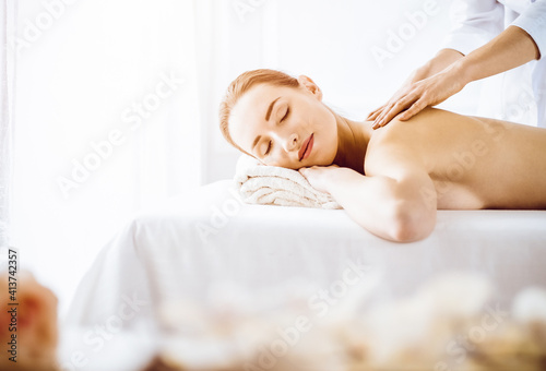 Beautiful woman enjoying back massage with closed eyes in sunny spa center. Relaxing treatment concept in medicine