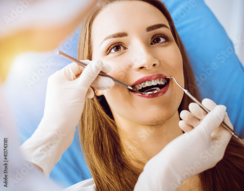 Smiling young woman with orthodontic brackets examined by dentist in sunny dental clinic. Healthy teeth and medicine concept