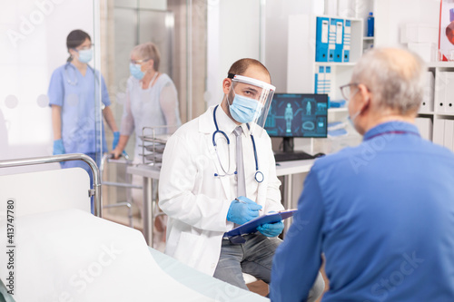 Doctor wearing visor as safety precaution in the course of consultation of senior man. Physician practitioner in the course of private modern clinic appointment during coronavirus pandemic.
