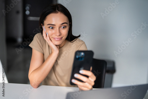 Young smiling cheerful woman indoors at home kitchen using social media apps on phone for video chatting and stying connected with her loved ones. Stay at home  social distancing lifestyle.