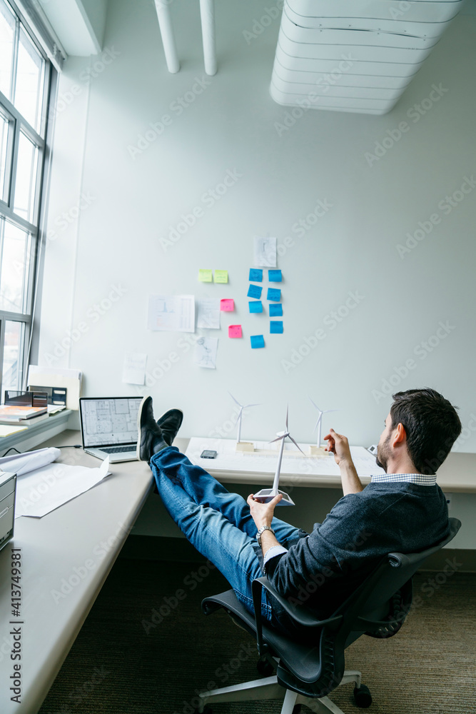 © Cavan Images - Side view of businessman holding wind turbine model while sitting in office © Cavan Images - Side view of businessman holding wind turbine model while sitting in office