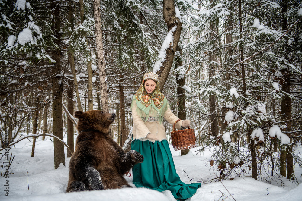 Russian beauty in folk national dress collects snowdrops in the winter forest with a brown bear