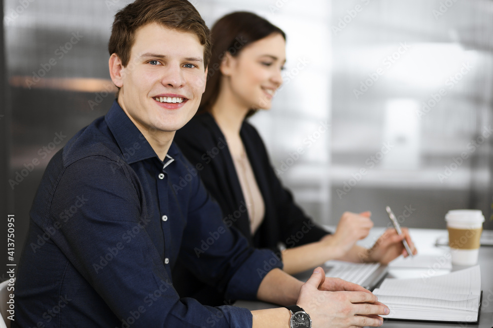 Handsome young businessman in a blue shirt is looking at camera with a smile, while he is sitting together with his female colleague at the desk in a cabinet. Focus on man. Headshot or business Handsome young businessman in a blue shirt is looking at camera with a smile, while he is sitting together with his female colleague at the desk in a cabinet. Focus on man. Headshot or business