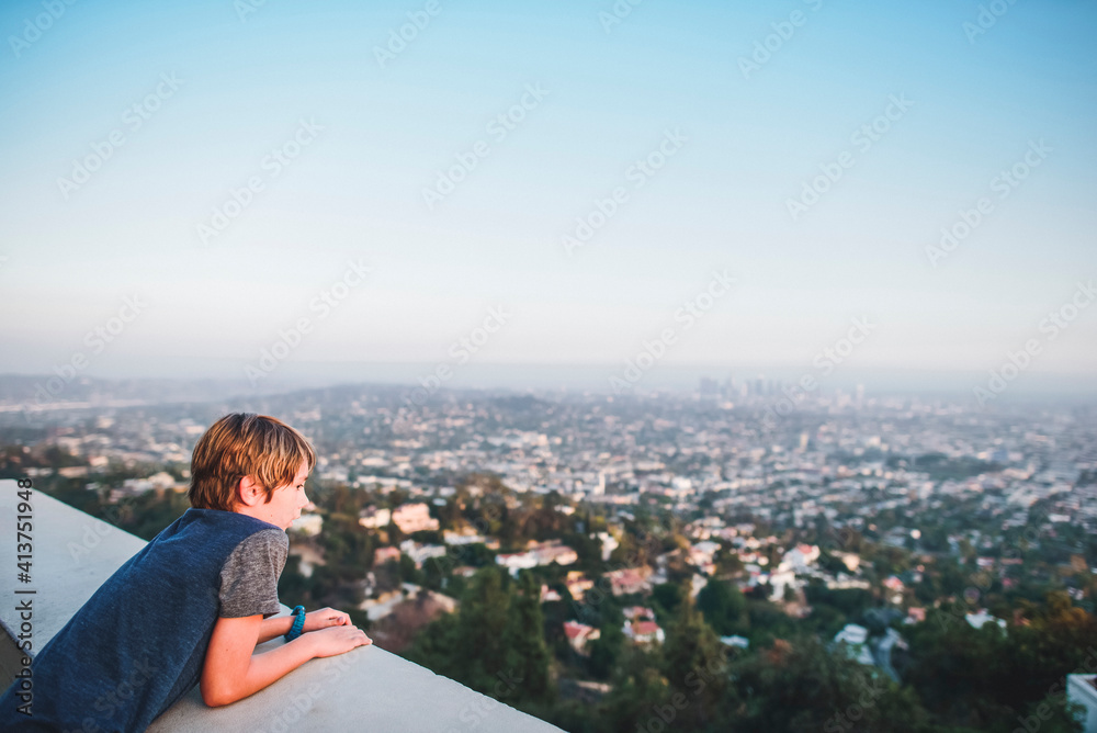 High angle view of boy looking at cityscape while leaning on retaining wall against sky at terrace High angle view of boy looking at cityscape while leaning on retaining wall against sky at terrace