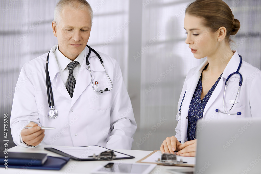 Doctors colleagues and elderly therapist discussing current disease therapy while sitting at working place in clinic. Teamwork in hospital. Medicine and healthcare concept Doctors colleagues and elderly therapist discussing current disease therapy while sitting at working place in clinic. Teamwork in hospital. Medicine and healthcare concept