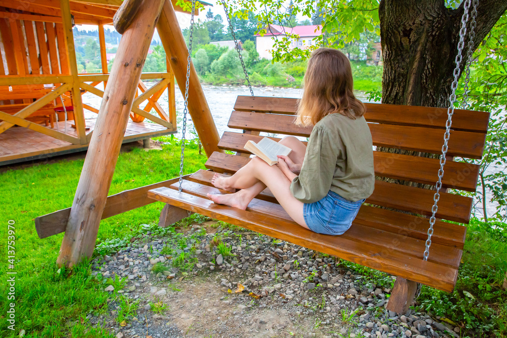 Hipster girl is reading a book on wooden bench. Woman relaxing outdoors on porch swing in the nature. Cozy photo. Summer mood. Hipster girl is reading a book on wooden bench. Woman relaxing outdoors on porch swing in the nature. Cozy photo. Summer mood.