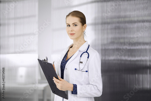 Cheerful female doctor standing in clinic. Portrait of friendly smiling woman physician. Perfect medical service in hospital. Medicine concept