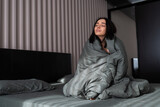 Attractive young woman is smiling and looking at the camera while sitting in bed under the blanket
