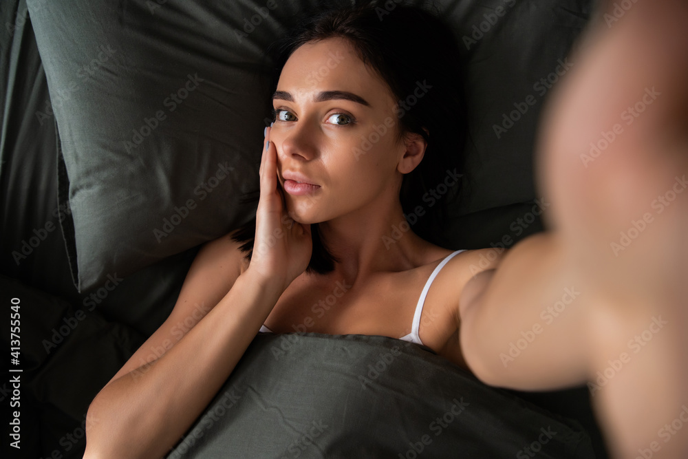 Obraz premium Self portrait of woman lying in bed shooting selfie on front camera enjoying recreation biting finger having online meeting with lover health healthy