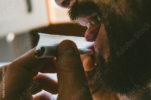 Bearded man licking end of rolling paper while making marijuana joints at home