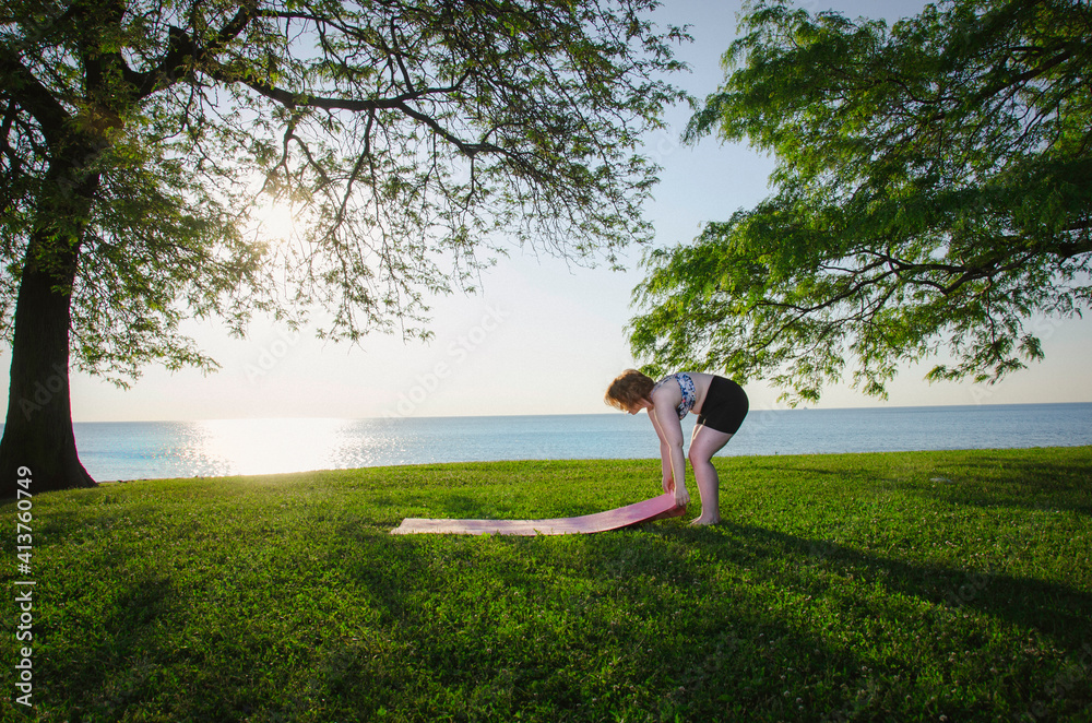 Woman adjusting exercise mat on grassy field at park against sea and clear sky Woman adjusting exercise mat on grassy field at park against sea and clear sky