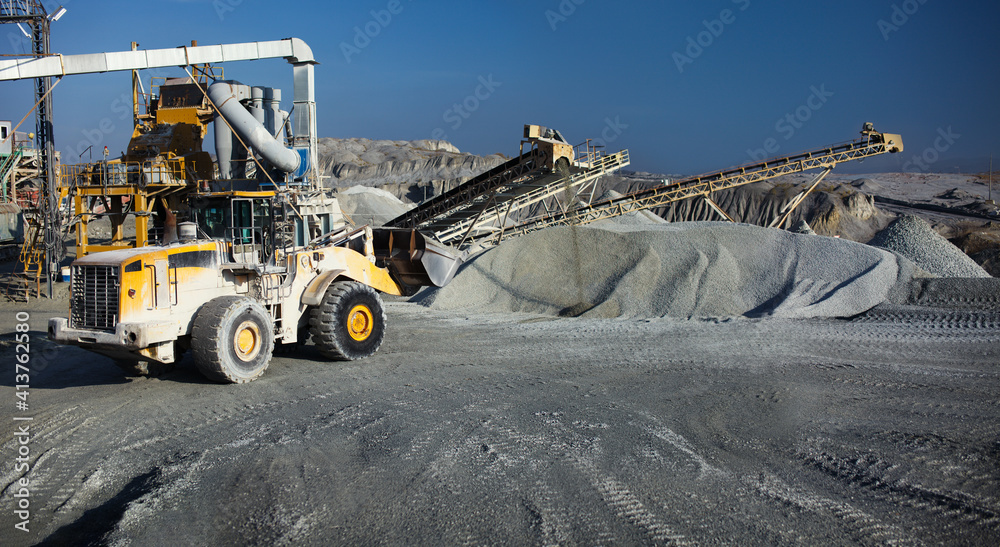 Panorama of rock stone crushing equipment at a mining factory with a front-end backhoe loader in the foreground.