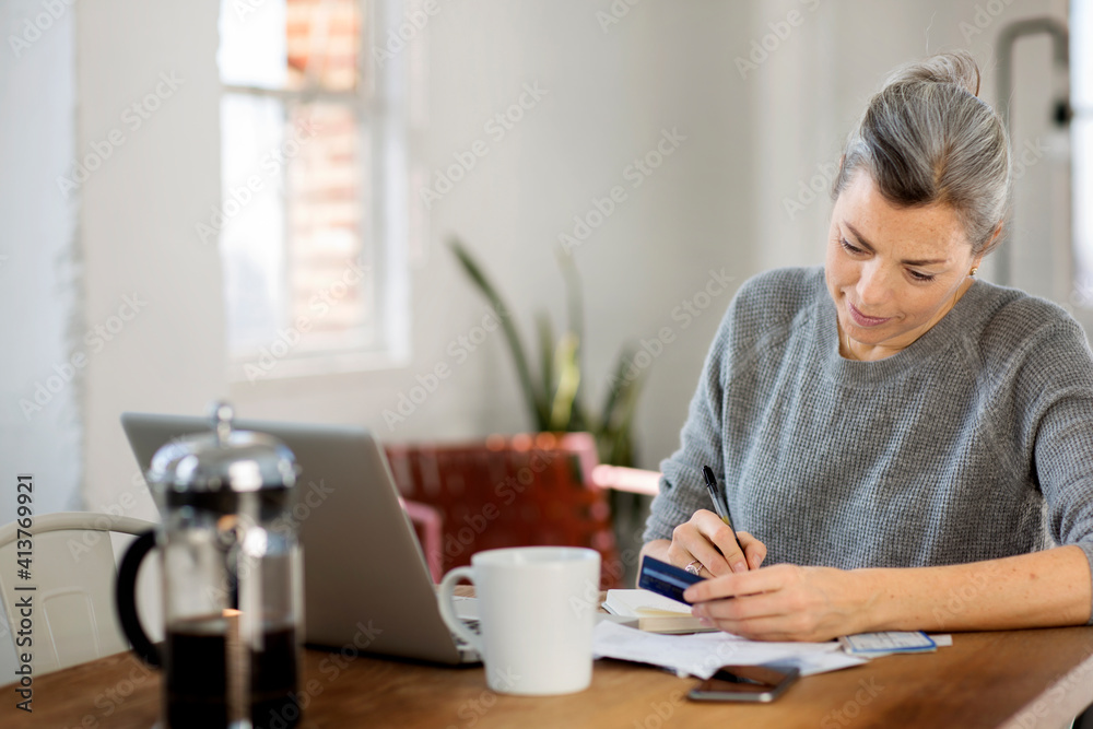 Mature woman writing in book while holding debit card at table Mature woman writing in book while holding debit card at table