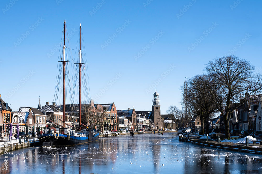 People skate on the ice in Lemmer with a ketch frozen in the ice on the left and the reformed church at the back. Sunny winter scene 