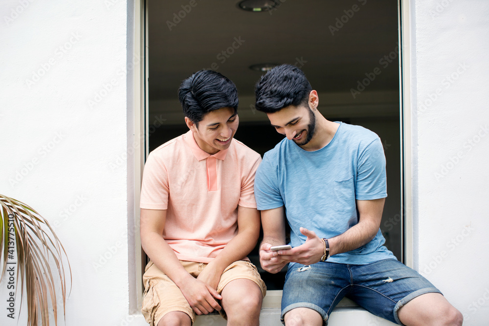 Happy male friends using smart phone while sitting on window