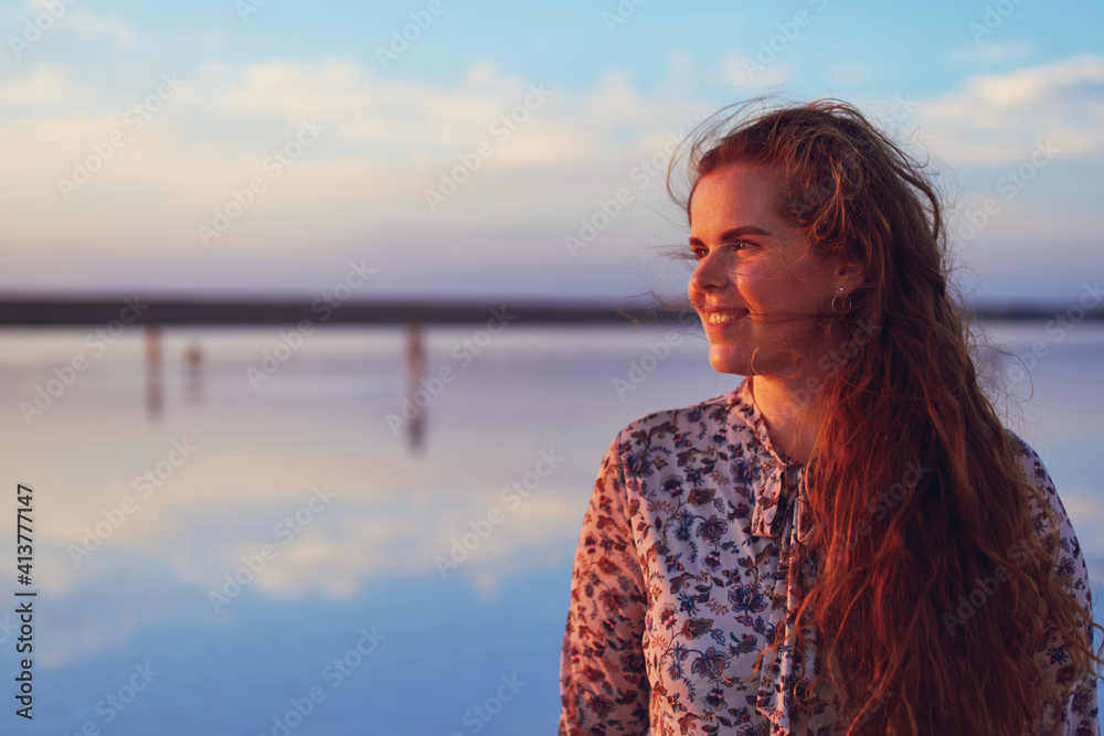 Beautiful young woman with flying red hair on the ocean coast at sunset time