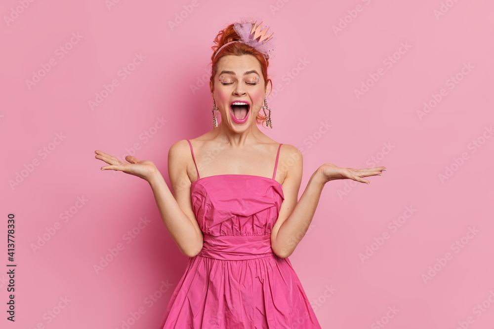 Emotional pretty redhead European lady spreads palms and exclaims loudly wears festive pink dress comes on party poses indoor. Birthday girl celebrates anniversary isolated over rosy background Emotional pretty redhead European lady spreads palms and exclaims loudly wears festive pink dress comes on party poses indoor. Birthday girl celebrates anniversary isolated over rosy background