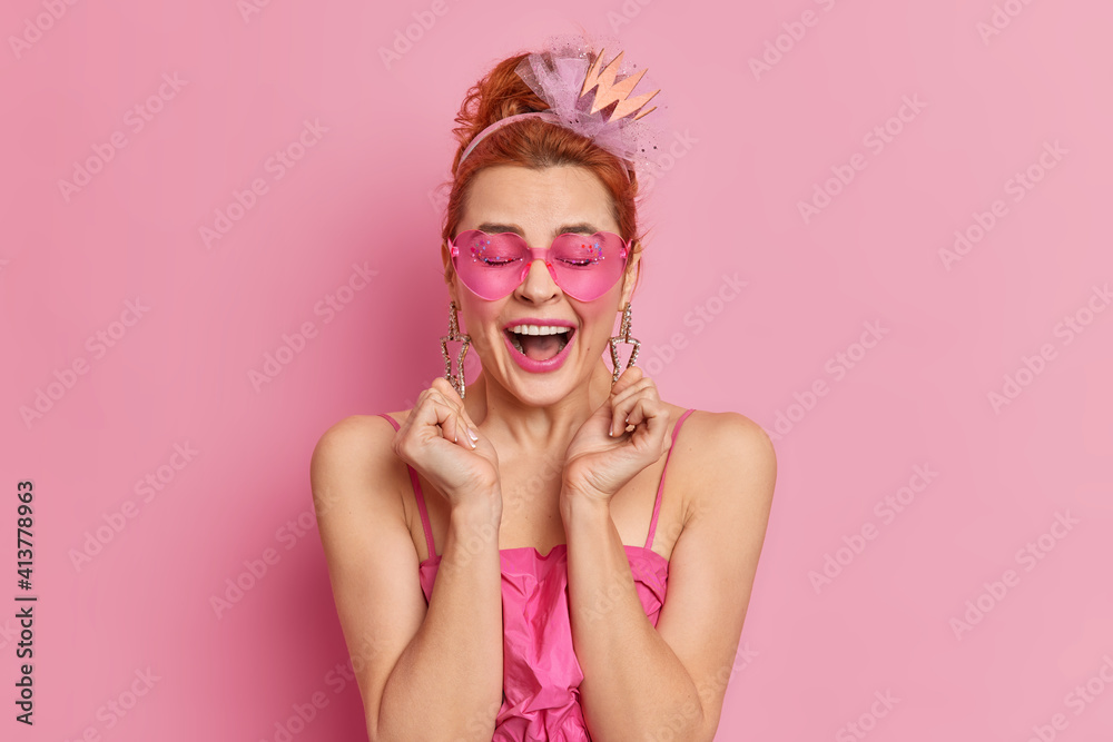 Overjoyed redhead young European woman raises hands wears trendy sunglasses festive dress and earrings expresses positive emotions isolated over pink studio background. People fashion style concept