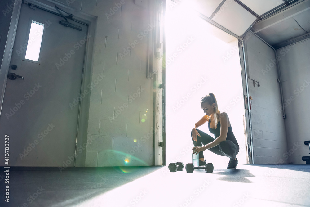 Tired woman holding water bottle while crouching in brightly lit gym Tired woman holding water bottle while crouching in brightly lit gym