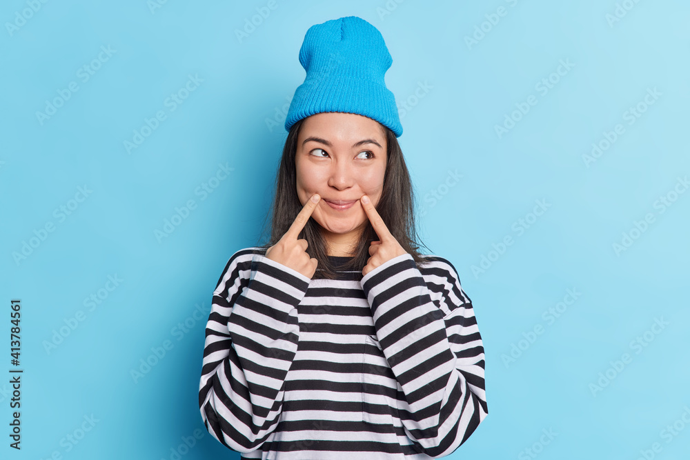 Asian woman with dark hair keeps fingers near corner of lips has satisfied expression looks aside dressed in long sleeved striped jumper and hat has fake smile on face isolated over blue background Asian woman with dark hair keeps fingers near corner of lips has satisfied expression looks aside dressed in long sleeved striped jumper and hat has fake smile on face isolated over blue background