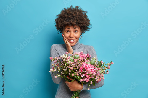 Portrait of cheerful African American woman accepts congratulations on birthday receives flowers has surprised expression dressed in casual turtleneck isolated over blue background. Special occasion
