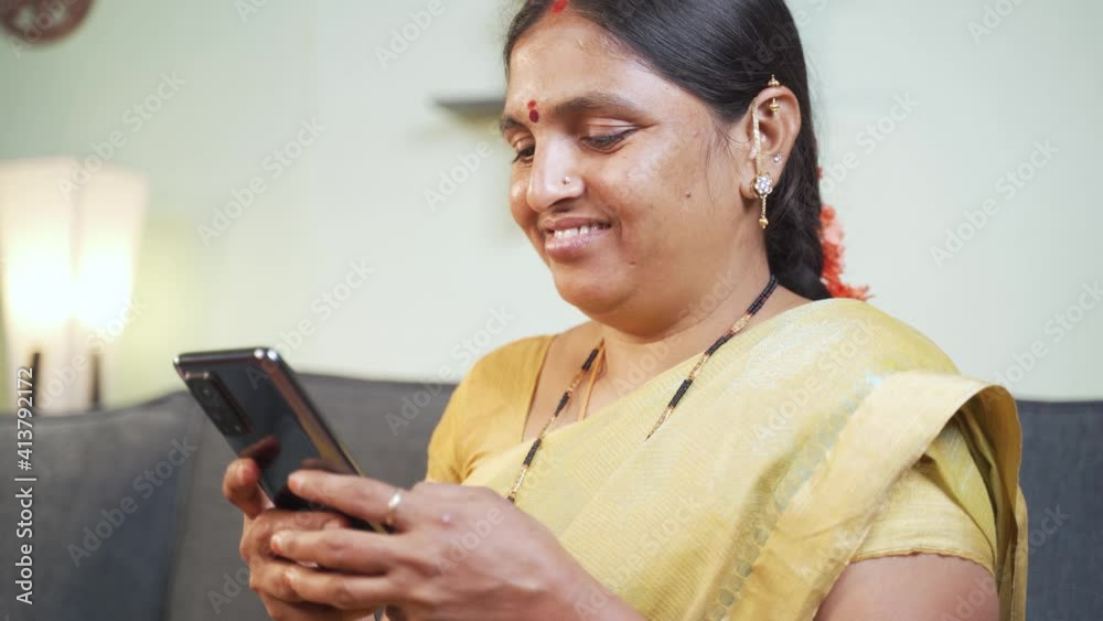 Indian woman busy using mobile phone while sitting on sofa - Concept of people using social media, online messaging app, Internet and entertainment lifestyle