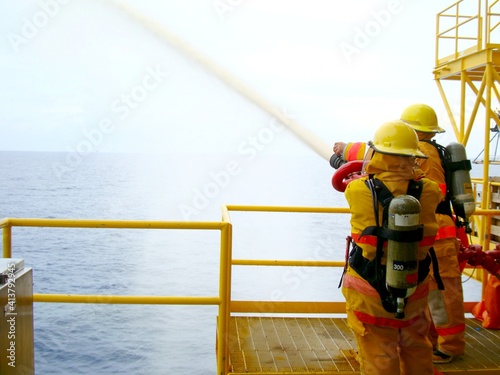Fire drill training on board for the crew in ship on offshore plant form oil and gas with fireman, fire hose, water spray, and blue sky, sea background.