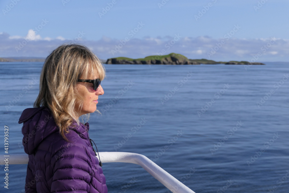An attractive blonde haired lady looks out to sea with sunglasses on a sunny day.Sea and island in background