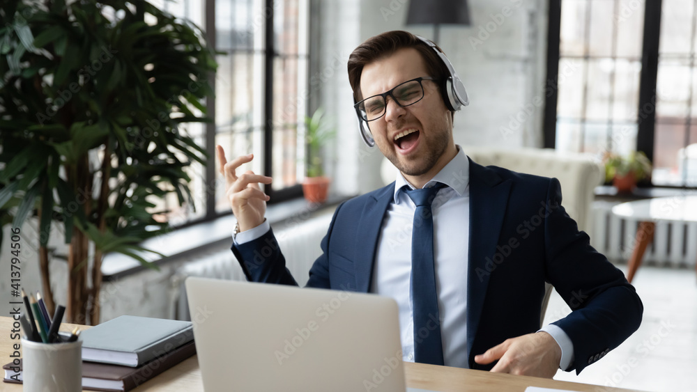 Fototapeta premium Overjoyed young Caucasian businessman in earphones sit at desk in office listen to music distracted from computer work. Happy male CEO in headphones have fun relax enjoy good quality sound in gadget.