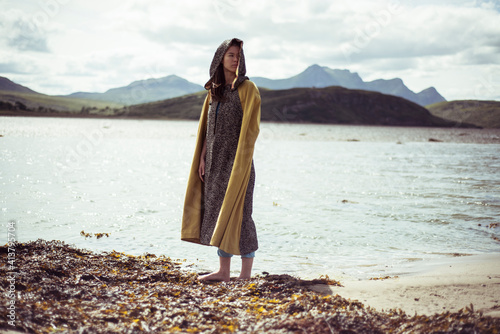 person in cloak stands on lake beach in mountains