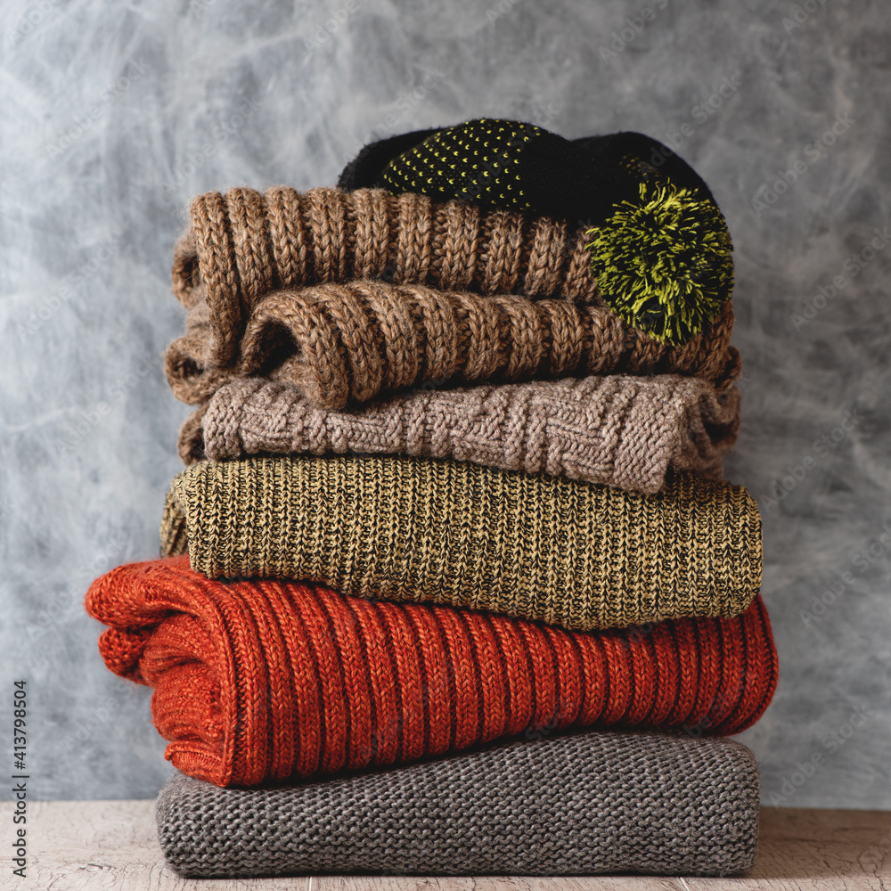 Stack of folded knitted wool women sweaters, scarf, hat with pompom on shelf against gray wall. Season of warm knitwear.