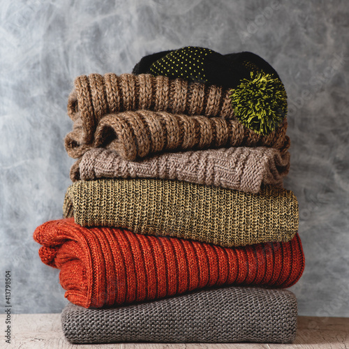 Obraz na płótnie Stack of folded knitted wool women sweaters, scarf, hat with pompom on shelf against gray wall. Season of warm knitwear. w salonie