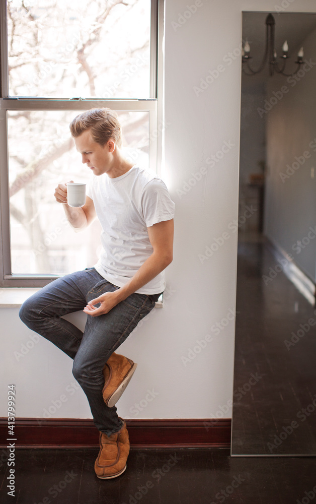 Man having drink while sitting on window sill at home Man having drink while sitting on window sill at home