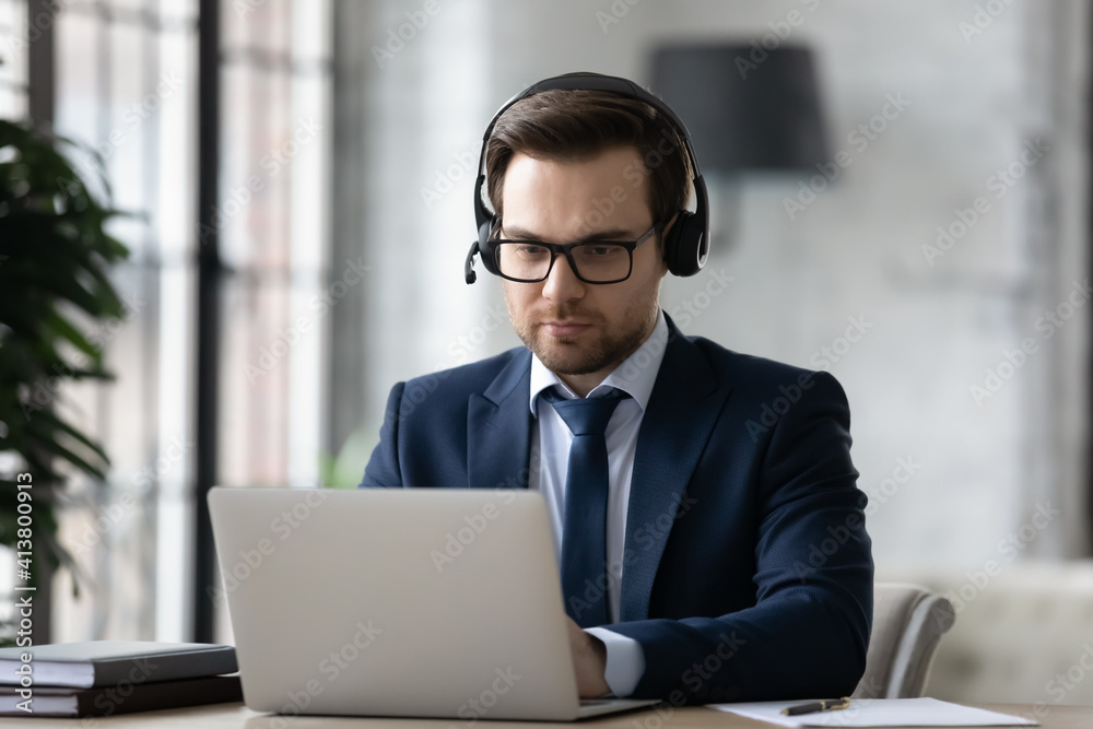Young Caucasian businessman in headphones sit at desk in office work talk online on video call on computer. Male CEO or employee in earphones have webcam virtual conference with partner on laptop. Young Caucasian businessman in headphones sit at desk in office work talk online on video call on computer. Male CEO or employee in earphones have webcam virtual conference with partner on laptop.