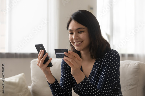 Smiling asian woman capable ebanking app user hold credit card smartphone satisfied with easy quick safe way to pay. Positive young lady sit on couch check bank account balance provide distant payment