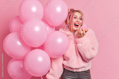 Positive young teenage girl with bright pink makeup bites delicious sweet doughnut dressed in coat going on birthday party holds inflated balloons isolated over rosy background. Festive concept