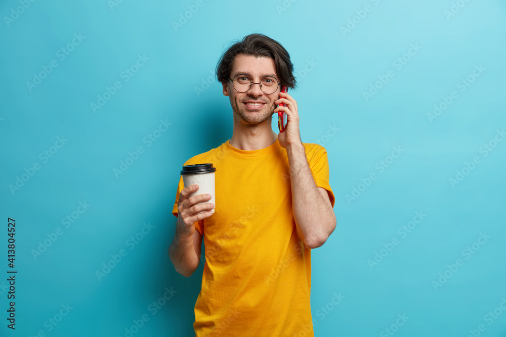 Photo of handsome adult European man has telephone conversation via smartphone drinks coffee to go wears round spectacles and yellow t shirt isolated over blue background enjoys good connection Photo of handsome adult European man has telephone conversation via smartphone drinks coffee to go wears round spectacles and yellow t shirt isolated over blue background enjoys good connection