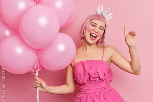 Joyful Asian woman tilts head raises arm feels very happy on party dressed in fashionable pink dress sings song along holds bunch of balloons poses indoor. People celebration entertainement and fun