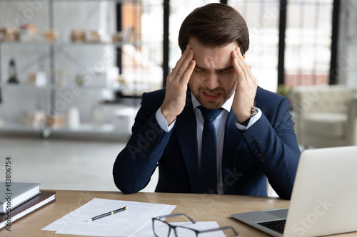 Unwell exhausted Caucasian young businessman sit in office working on computer massage temples suffer from headache. Tired unhealthy male CEO struggle with migraine, overwhelmed with screen job.