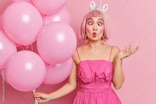 Beautiful young Asian woman makes fish lips has funny grimace raises hand wears bright makeup raises hand holds helium balloons isolated over pink background celebrates anniversary or birthday