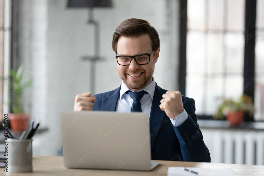 Overjoyed young Caucasian businessman sit at desk in office look at computer screen triumph win lottery online. Happy male boss use laptop read good business news. Financial success concept. Overjoyed young Caucasian businessman sit at desk in office look at computer screen triumph win lottery online. Happy male boss use laptop read good business news. Financial success concept.