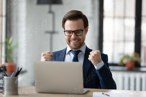 Overjoyed young Caucasian businessman sit at desk in office look at computer screen triumph win lottery online. Happy male boss use laptop read good business news. Financial success concept.