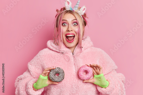 Positive cheerful Caucasian woman laughs happily keeps mouth opened holds glazed doughnuts dressed in fur coat isolated over pink background feels very happy. Funny millennial girl has fun with donuts