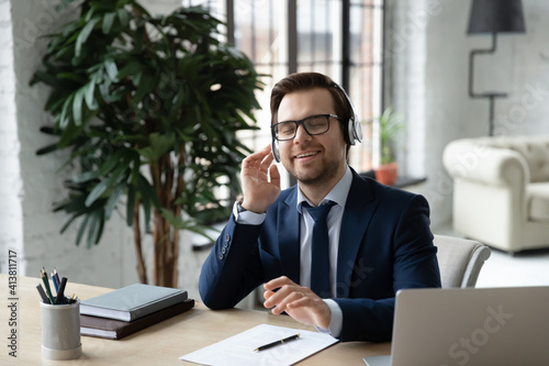 Happy male CEO or boss sit relax at office desk listen to music in modern headphones. Smiling young Caucasian businessman in earphones enjoy good quality sound on gadget. Technology concept.
