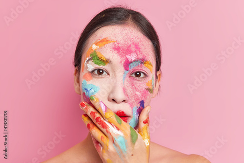 Close up shot of young female fashion model has vivid paint makeup bright colors holds chin smeared with oil paints isolated over pink background. Art and creativity concept. Abstract makeup