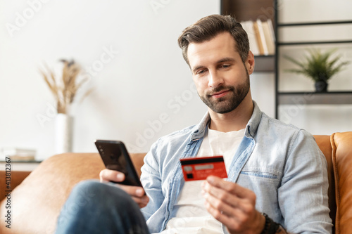 Bearded handsome man makes purchase online. A hipster wearing casual shirt holds a mobile phone and a credit card orders food. Smiling guy is using smartphone and debit bank card for paying in e-shop photo