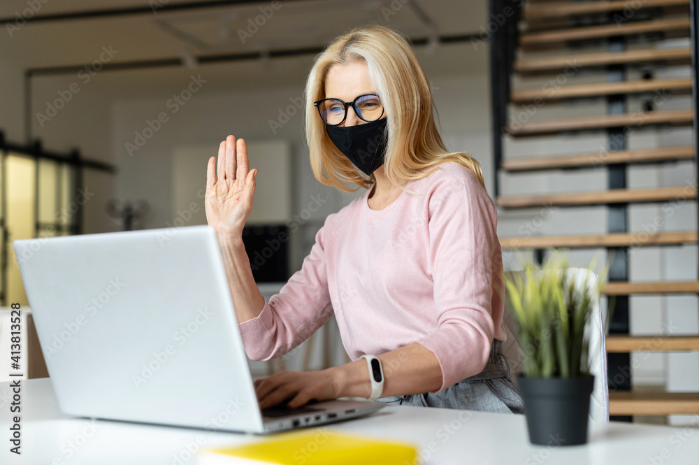 Elegant middle-aged woman sitting at the laptop in the office during a pandemic,greetings colleagues,chatting with friends,smiling, gives the high-five,looking at laptop screen,on an online meeting